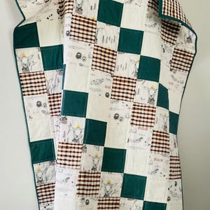 May include: A patchwork quilt featuring a repeating pattern of white, green, and brown squares. Some squares have a gingham pattern, others have illustrations of goats wearing crowns. The quilt has a green border.