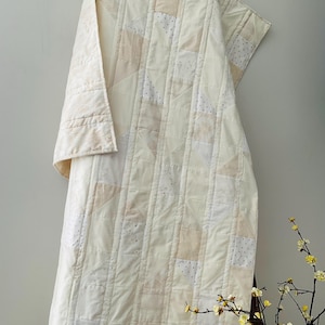 May include: A white and cream patchwork quilt with a subtle pattern of small dots. The quilt is folded and draped over a wooden ladder.