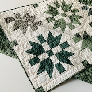 May include: A handmade quilt with a white background and green star patterns. The quilt features a mix of solid and patterned fabrics in shades of green and white. The edges are finished with a green floral fabric.