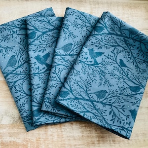 May include: Four folded cloth napkins with a teal bird and tree branch pattern on a light blue background. The napkins are made of fabric and are arranged in a stack. The design features birds perched on branches.