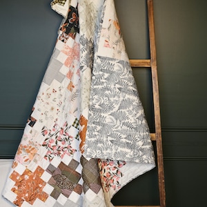 May include: A patchwork quilt with various floral and geometric patterns, draped over a wooden ladder. The quilt features a mix of colors including white, pink, brown, and green. The quilt's design includes floral prints and cross-stitch patterns.