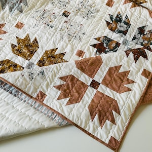 May include: A quilted blanket featuring a repeating bear paw block pattern in shades of terracotta, mustard yellow, and cream. The fabric includes floral and speckled prints, with white background fabric and straight-line machine quilting throughout.