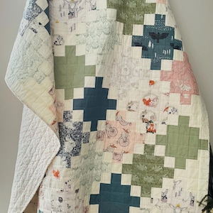 May include: A handmade quilt with a patchwork design. The quilt features a variety of patterns, including floral, woodland creatures, and geometric shapes. The color palette includes pink, green, blue, and cream. The quilt is approximately 70 inches by 80 inches.