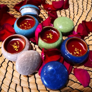 May include: Six small, round, ceramic containers with lids, filled with a red substance and gold flakes. The containers are arranged on a woven mat with rose petals scattered around them.