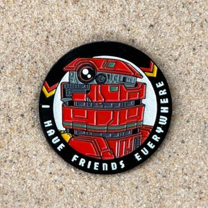 May include: A circular enamel pin featuring a red robotic character with a single large eye. The background is black with white text that reads I HAVE FRIENDS EVERYWHERE, accented by two yellow and red chevron symbols on the left and right sides.