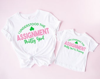 May include: Two white t-shirts with pink and green text. The larger shirt says "I understood the assignment Pretty Girl" and the smaller shirt says "I'm learning the assignment Pretty Girl in training".