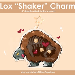 Könnte beinhalten: Ein 7,6 cm großer doppelseitiger Shaker-Anhänger mit einem Lox-Cartoon, einer mythischen Kreatur mit einem pelzigen braunen Körper und einem grauen, schuppigen Kopf. Der Lox hält ein rotes Herz und es befinden sich weitere Herzen und weiße Perlen im Anhänger.