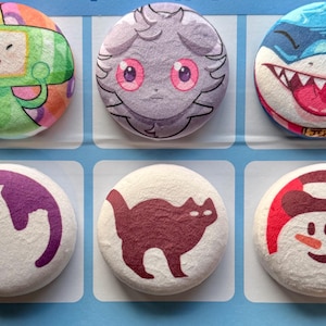 May include: Six circular badges with cartoon illustrations. The designs include a robot, a creature with pink eyes, a shark, a ghost, a cat, and a snowman. The badges are brightly coloured and set against a light blue background.