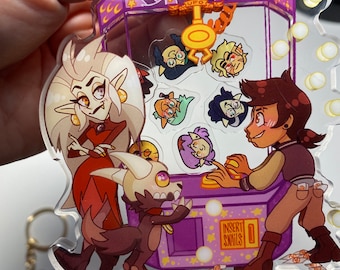 May include: A clear acrylic keychain featuring characters from The Owl House animated series. The keychain depicts Eda, King, Luz, and a claw machine with the text "The Collector". The claw machine has the text "Insert Snails".