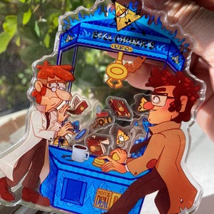 May include: A clear acrylic keychain featuring a blue claw machine with two cartoon characters. The machine has the text "UFO" and "Insert Deal". The characters are in brown, red, and white clothing.