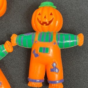 May include: A plastic scarecrow figurine with a carved pumpkin head, wearing orange overalls with purple striped sleeves and purple patches. The figure has a small green hat on top of the pumpkin head and features a smiling jack-o'-lantern face.