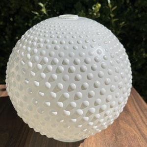 May include: A spherical white glass lamp shade featuring a raised hobnail texture across its entire surface. The globe has a circular opening at the top and a smooth, flat base, showcasing a consistent opaque finish with a repeating pattern of small, rounded bumps.