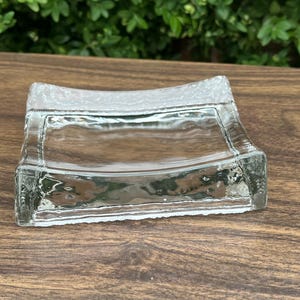 May include: A square clear glass dish with a concave centre and a textured, frosted border. The dish features thick, curved edges and a smooth, transparent basin, creating a simple and functional design for holding small items or decorative objects.