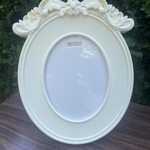 May include: An oval-shaped picture frame in a solid off-white color. The frame features a smooth, rounded inner border and an ornate, decorative top section with sculpted floral and leaf-like patterns. The center contains a blank white insert.