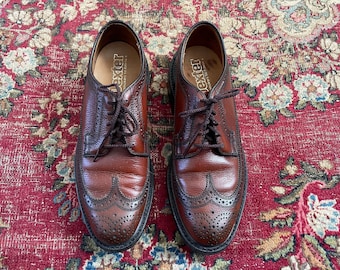 May include: A pair of brown leather brogue shoes with wingtip detailing and laces. The shoes are on a red patterned rug.