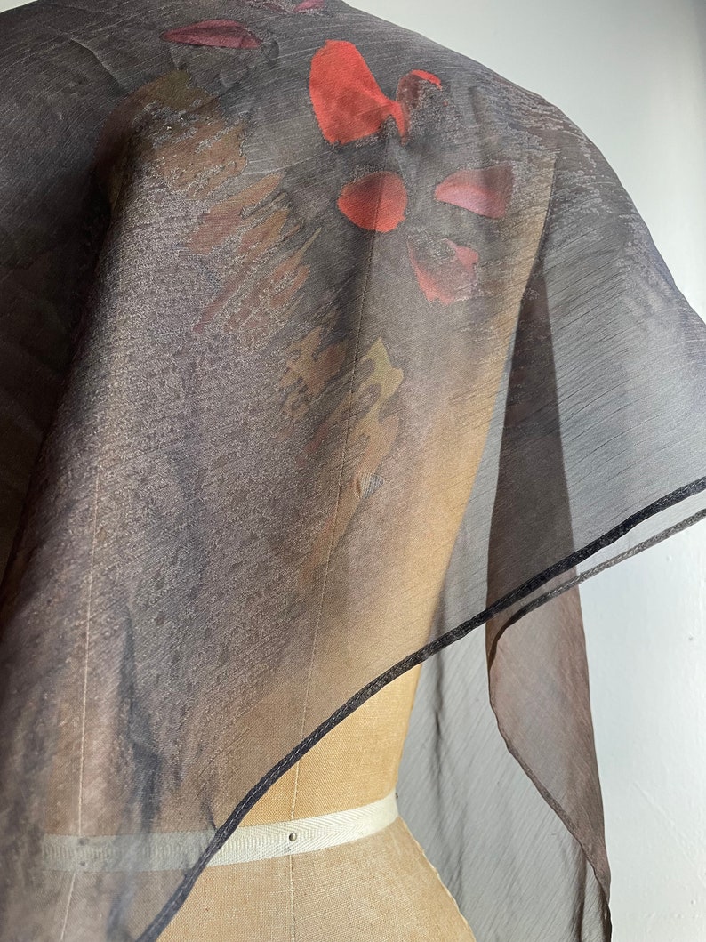 May include: A sheer black scarf with a floral print in shades of red, orange, and purple. The scarf is trimmed with a black border.