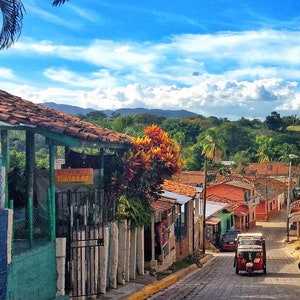 Puede incluir: Una escena de calle colorida en un pueblo peque&ntilde;o con un veh&iacute;culo de tres ruedas rojo y negro estacionado en la calle. El veh&iacute;culo tiene un letrero que dice "Souvenirs Tortillas". La calle est&aacute; bordeada de edificios coloridos con techos de terracota.
