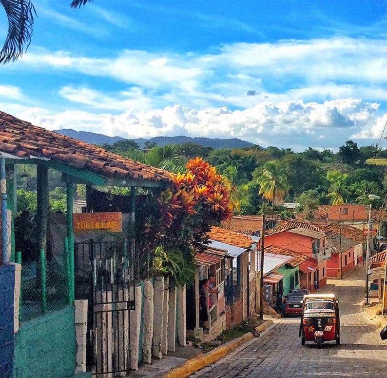 Puede incluir: Una escena de calle colorida en un pueblo peque&ntilde;o con un veh&iacute;culo de tres ruedas rojo y negro estacionado en la calle. El veh&iacute;culo tiene un letrero que dice "Souvenirs Tortillas". La calle est&aacute; bordeada de edificios coloridos con techos de terracota.
