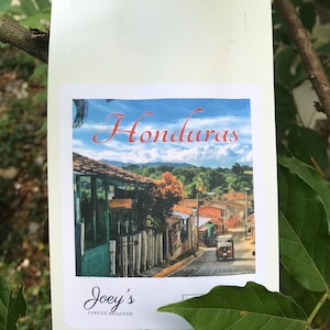 Puede incluir: Una bolsa blanca de granos de caf&eacute; con una etiqueta que dice "Honduras" y muestra una foto de una escena callejera en una ciudad. La etiqueta tambi&eacute;n dice "Joey's Coffee Roaster".