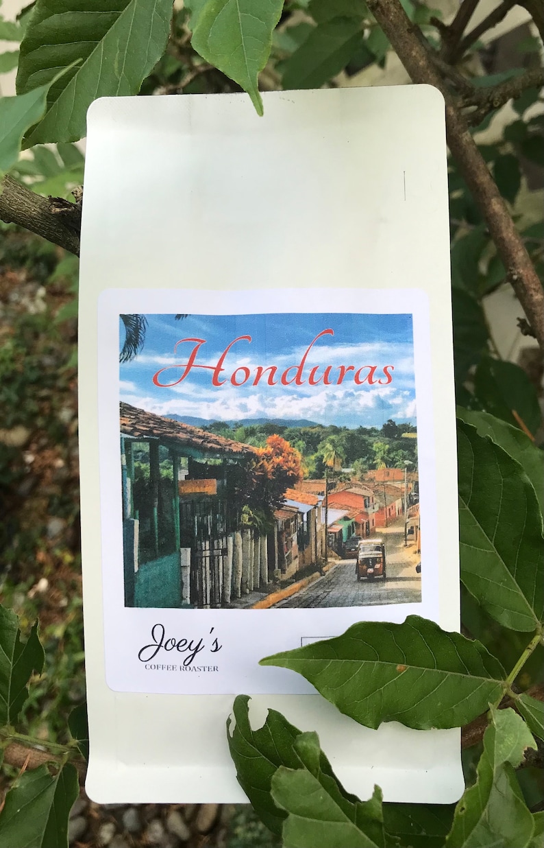 Puede incluir: Una bolsa blanca de granos de caf&eacute; con una etiqueta que dice "Honduras" y muestra una foto de una escena callejera en una ciudad. La etiqueta tambi&eacute;n dice "Joey's Coffee Roaster".