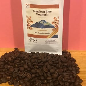 May include: A white bag of Joey's Jamaican Blue Mountain coffee with a mountain graphic. Dark roasted coffee beans are scattered in front of the bag. The bag reads "Premium Grade" and "Blue Mountains, Jamaica."