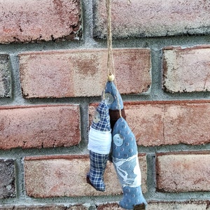 May include: Three blue and white fabric fish ornaments hanging from a string. The fish are decorated with buttons and have a rustic, handmade look.