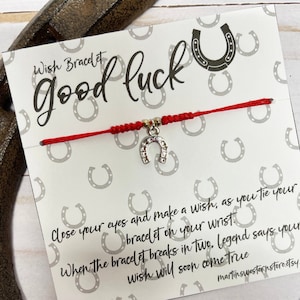 Puede incluir: Una pulsera roja de deseos con un dije de herradura plateada. La tarjeta dice "Wish Bracelet Good Luck" con instrucciones y un sitio web. La pulsera se muestra cerca de una herradura.