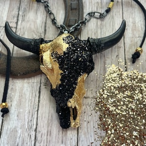 May include: A gold and black glitter bull skull pendant necklace with a black leather cord. The pendant is made of resin and has a detailed skull design.
