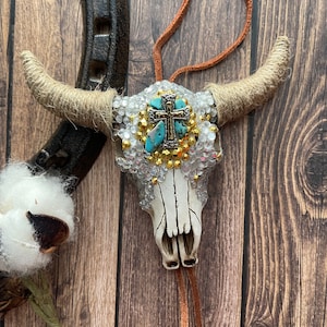 May include: White cow skull with a turquoise and gold cross on the forehead. The skull is decorated with rhinestones and has a brown leather cord hanging from the bottom.