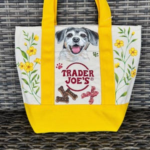 May include: A cream-coloured canvas tote bag with bright yellow handles and a yellow base. The bag features a central illustration of a dog, the red Trader Joe's logo, dog treats, and bacon, flanked by yellow floral stems on both sides.