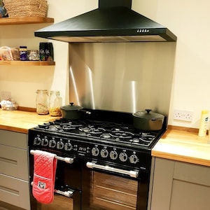 May include: Black range cooker with a stainless steel splashback and a black range hood above it. The cooker has a red tea towel hanging on the handle.