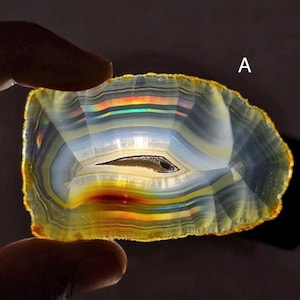 May include: A translucent, polished agate slice with a central eye-like pattern. The stone features layers of color, including yellow, orange, blue, and gray, with a rainbow effect. The letter "A" is visible in the upper right corner.