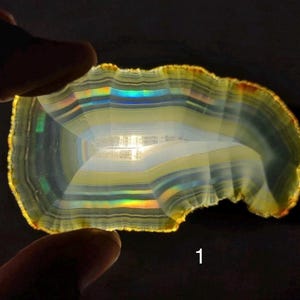 May include: A translucent, layered agate slice with a vibrant display of colors. The stone features bands of yellow, white, and iridescent hues, with a textured edge. The agate is held between two fingers against a dark background, with the number "1" visible.