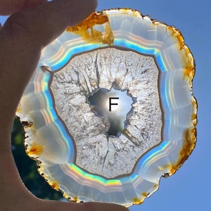 May include: A translucent, circular agate slice with a central crystalline geode cavity marked with the letter F. The stone features concentric white and iridescent bands with amber-colored edges, backlit to reveal internal mineral textures.