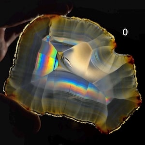 May include: A translucent, polished gemstone slice with a rainbow spectrum of colors. The stone has a layered appearance with a light brown and yellow outer edge. The number "0" is visible in the upper right corner.