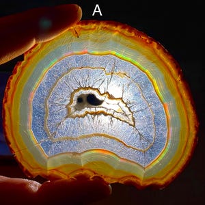 May include: A translucent agate slice labeled with the letter A, featuring concentric bands of orange, yellow, and white. The center is a hollow, crystalline cavity that glows with light, revealing intricate internal fractures and iridescent colors.