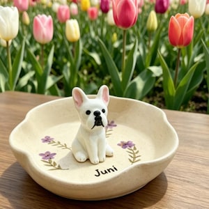 May include: A cream-colored, scalloped-edge dish with a white French Bulldog figurine. The dish is decorated with purple floral accents and the name "Juni" in black script. The background features a vibrant garden of colorful tulips.