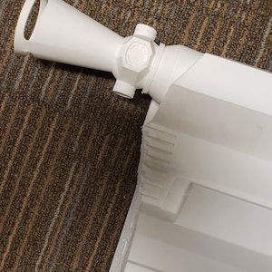 May include: A white, 3D-printed rifle scope with a cylindrical body and a circular sight. The scope has a handle and several adjustment knobs. The object is set against a brown and tan patterned surface.