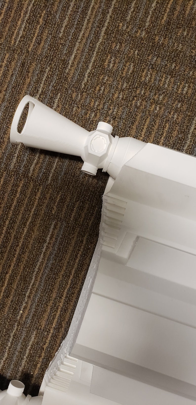 May include: A white, 3D-printed rifle scope with a cylindrical body and a circular sight. The scope has a handle and several adjustment knobs. The object is set against a brown and tan patterned surface.