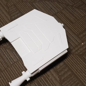 May include: A white, geometric-shaped object with a series of rectangular and angled panels. The object has two cylindrical extensions on either side. The item is made of a solid, opaque material.