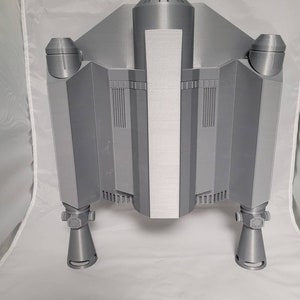 May include: A gray, 3D-printed model of a spaceship, featuring a central body with a vertical silver panel, flanked by angular wings and cylindrical engine components. The model has detailed vents and nozzles.