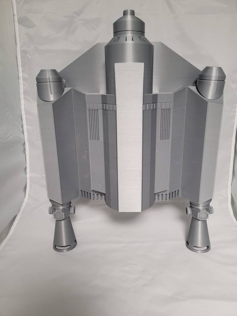 May include: A gray, 3D-printed model of a spaceship, featuring a central body with a vertical silver panel, flanked by angular wings and cylindrical engine components. The model has detailed vents and nozzles.