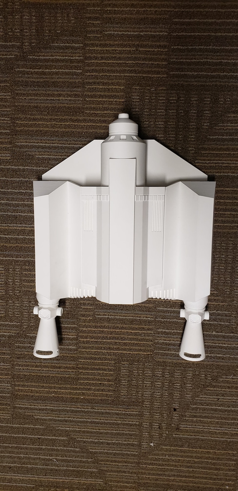 May include: A white, geometric model of a spacecraft, featuring a central body with angular wings and two lower engine pods. The model has a detailed design with vertical lines and a cylindrical top. The object is set against a brown textured surface.