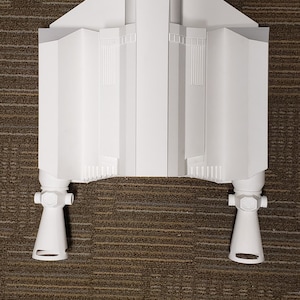 May include: A white model of a spacecraft, featuring a central body with angled wings and two lower engine components. The model is set against a brown textured background.