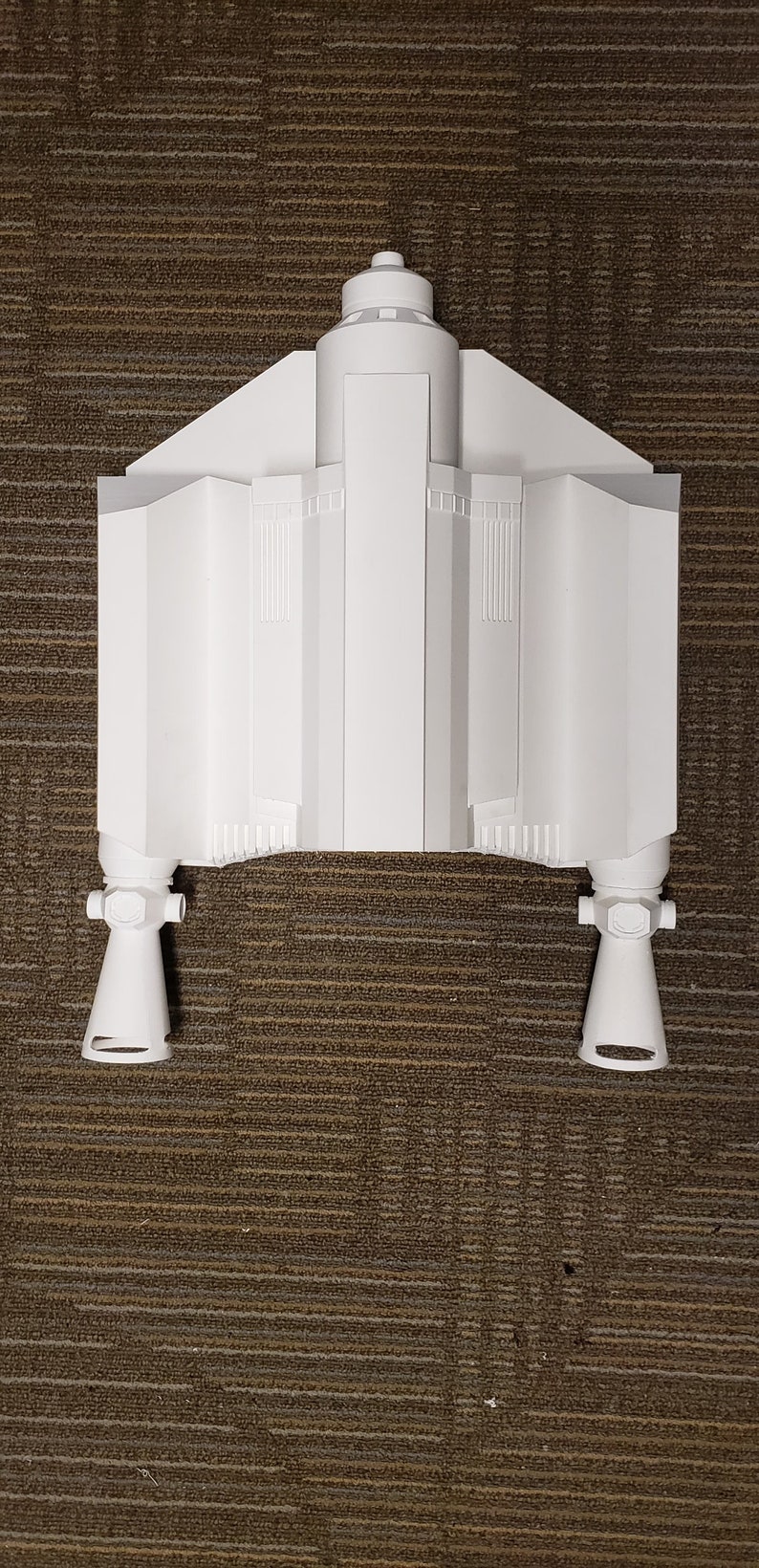 May include: A white model of a spacecraft, featuring a central body with angled wings and two lower engine components. The model is set against a brown textured background.