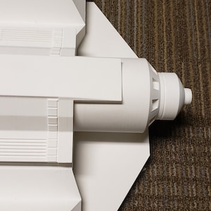 May include: A white model of a spacecraft, featuring a cylindrical body and angular wings. The model has detailed features, including ridges and a nozzle at the end. The spacecraft is set against a brown carpet.