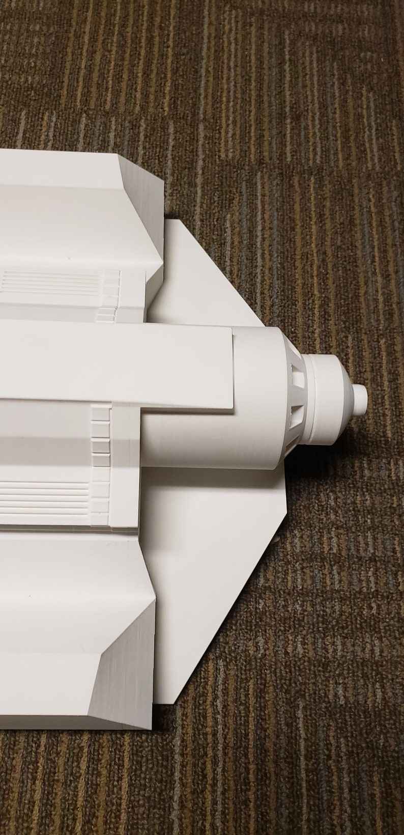 May include: A white model of a spacecraft, featuring a cylindrical body and angular wings. The model has detailed features, including ridges and a nozzle at the end. The spacecraft is set against a brown carpet.