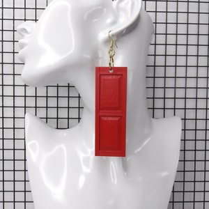May include: Red wooden door-shaped earrings with gold hooks. The earrings are long and rectangular.