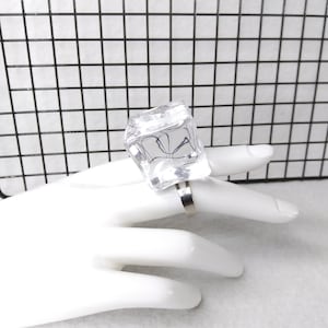 May include: A clear, square-shaped ring with a silver band. The ring is on a white mannequin hand.