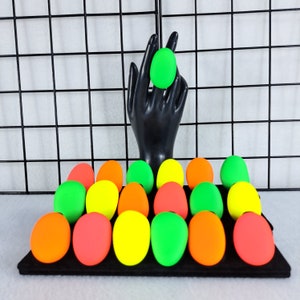 May include: A collection of 18 brightly colored, soft, squishy, egg-shaped stress balls in green, yellow, and orange. The stress balls are arranged in a semi-circle on a black surface.